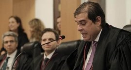 Advogado Paulo Cesar Salomão discursa ao microfone; ele está sentado, e veste toga; ao lado memb...