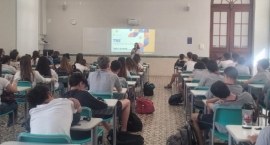 Uma sala de aula cheia de estudantes, sentados em carteiras verdes, assistindo a uma palestra mi...