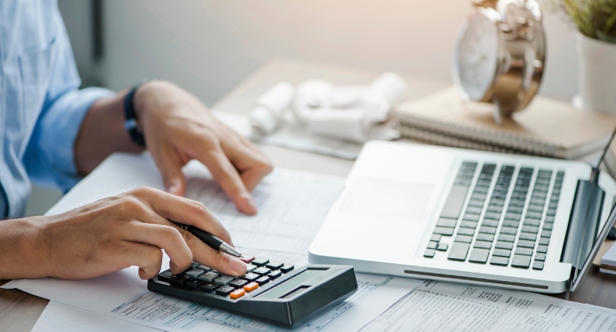 Pessoa trabalhando em uma mesa de escritório repleta de documentos financeiros. Com uma das mãos...