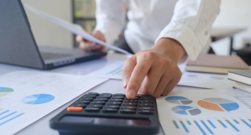 Pessoa trabalhando em uma mesa de escritório repleta de documentos financeiros. Com uma das mãos...