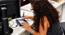 Mulher sentada diante de um computador utilizando uma mesa digitalizadora/caneta eletrônica para...