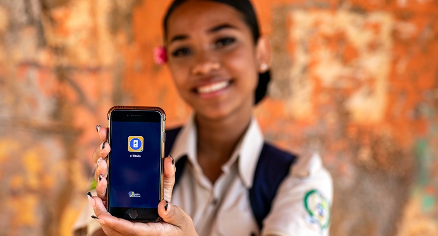 Jovem sorridente em uniforme escolar segura um celular em primeiro plano, exibindo a tela com o ...