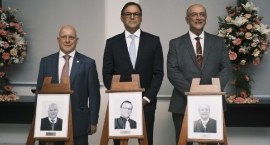 A imagem mostra três homens de terno e gravata posando para uma foto em um ambiente formal, poss...