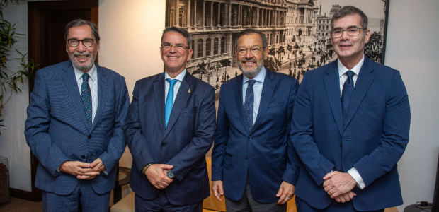 Quatro homens de terno e gravata posam lado a lado para uma foto, em ambiente interno, diante de...