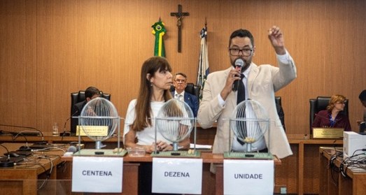 A imagem mostra um homem de óculos, vestindo terno claro, camisa branca e gravata escura, que se...