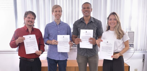 Foto posada dos quatro novos servidores do TRE-RJ; todos seguram o termo de posse. Da esquerda p...