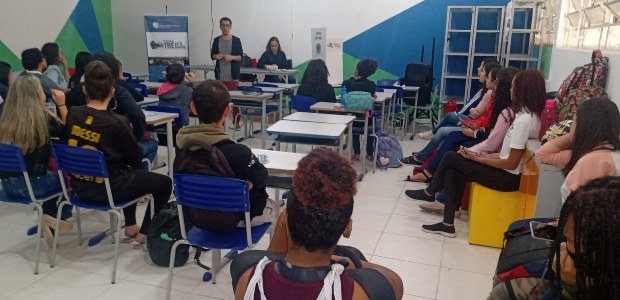 Na sala de aula, há várias alunas e alunos sentados. Em pé no fundo da sala, o servidor da EJE-R...