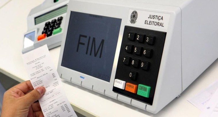TSE - Urna eletrônica modelo 2020 - Destaque Pessoa segurando o boletim de urna, em frente a urna eletrônica. Na tela da urna, está escrito FIM.