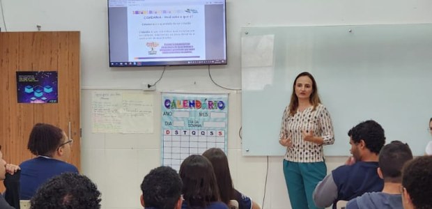 A imagem mostra uma sala de aula com estudantes sentados, olhando para a palestrante, que está p...