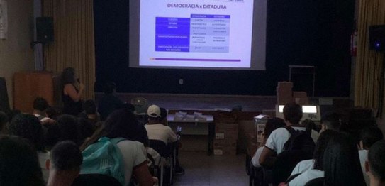 A imagem mostra uma sala de aula com um palestrante na frente, apresentando gráficos em uma tela...