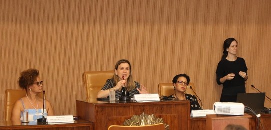 Três mulheres estão sentadas atrás de uma mesa de madeira e uma está de pé. O fundo é de madeira...