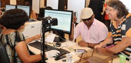 A foto mostra a interação entre eleitor e atendente durante um atendimento do TRE-RJ. Uma mesa s...