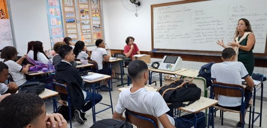 Sala de aula com estudantes sentados em cadeiras azuis assistindo a uma apresentação conduzida p...