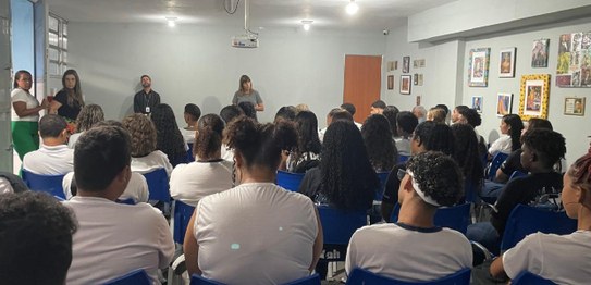 A imagem mostra um ambiente de sala de aula com um quadro branco ao fundo e estudantes sentados....