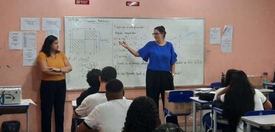 A imagem mostra um ambiente de sala de aula com um quadro branco ao fundo e estudantes sentados....