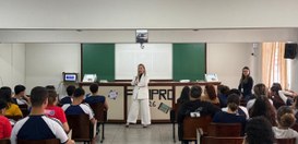 Uma mulher de terno branco palestra ao microfone em uma sala de aula de frente para uma plateia ...