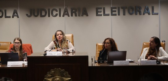 A imagem mostra uma longa mesa de madeira escura onde quatro mulheres estão sentadas lado a lado...