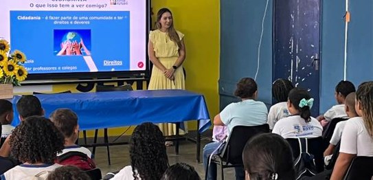 Sala de aula com bandeirinhas coloridas no teto; estudantes estão sentados assistindo a palestra
