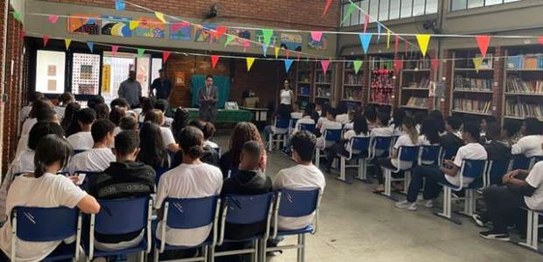 Sala de aula com bandeirinhas coloridas no teto; estudantes estão sentados assistindo a palestra
