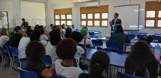 Sala de aula com bandeirinhas coloridas no teto; estudantes estão sentados assistindo a palestra

