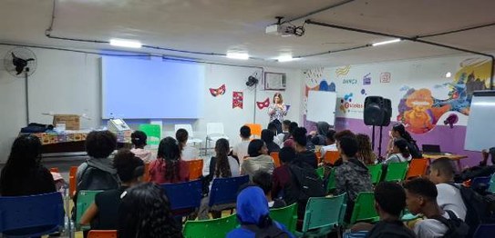 Sala de aula com bandeirinhas coloridas no teto; estudantes estão sentados assistindo a palestra
