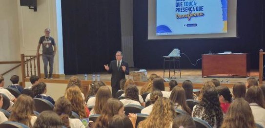 Sala de aula com bandeirinhas coloridas no teto; estudantes estão sentados assistindo a palestra
