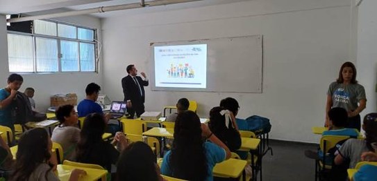Sala de aula com bandeirinhas coloridas no teto; estudantes estão sentados assistindo a palestra
