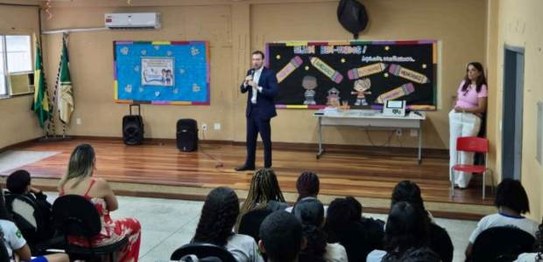 Sala de aula com bandeirinhas coloridas no teto; estudantes estão sentados assistindo a palestra
