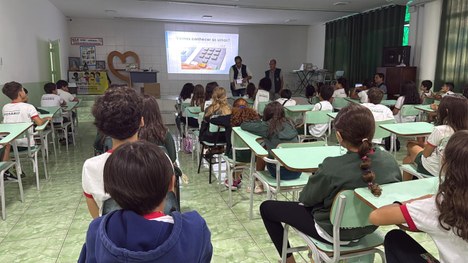 Sala de aula com bandeirinhas coloridas no teto; estudantes estão sentados assistindo a palestra
