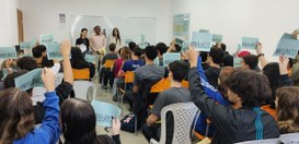 Alunos sentados em uma sala de aula levantam cartazes verdes com a palavra "CONFIRMA" escrita em...