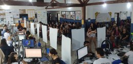 A imagem mostra uma sala ampla e movimentada, onde está sendo realizado atendimento eleitoral. D...