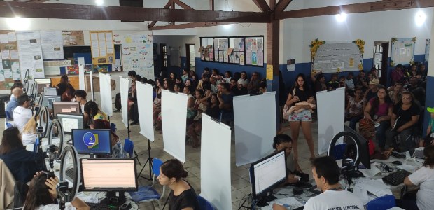A imagem mostra uma sala ampla e movimentada, onde está sendo realizado atendimento eleitoral. D...