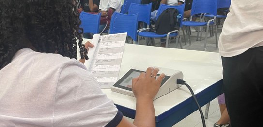 A imagem mostra um ambiente de sala de aula com um quadro branco ao fundo e estudantes sentados....