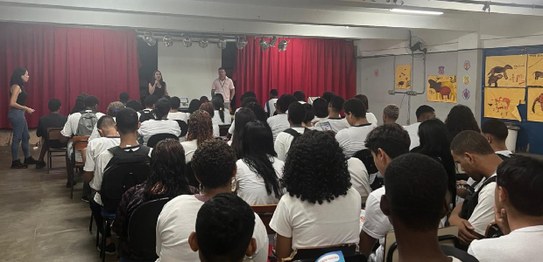 A imagem mostra um ambiente de sala de aula com um quadro branco ao fundo e estudantes sentados....