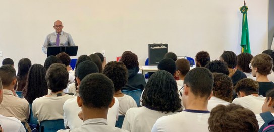 A imagem mostra um ambiente de sala de aula com um quadro branco ao fundo e estudantes sentados....