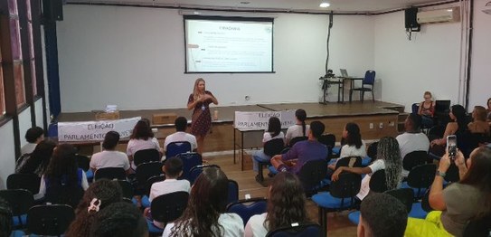 Dois homens falam para um grupo de crianças em uma sala de aula. Uma urna eletrônica está posici...