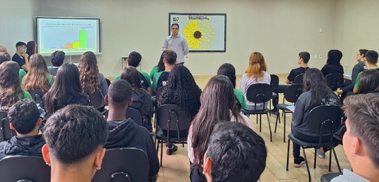 A imagem mostra uma sala de aula com um palestrante na frente, apresentando gráficos em uma tela...