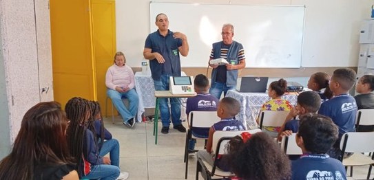 Dois homens falam para um grupo de crianças em uma sala de aula. Uma urna eletrônica está posici...