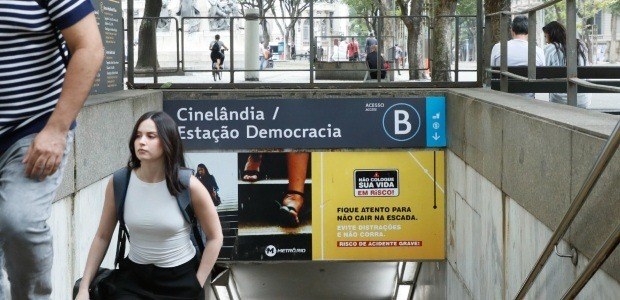 Entrada da estação de metrô vista da calçada. Uma mulher de regata branca e mochila caminha subi...