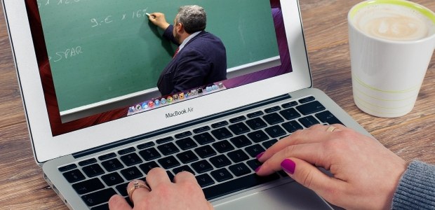 sobre uma mesa de madeira, há um laptop que mostra na tela um professor de terno e gravata escre...