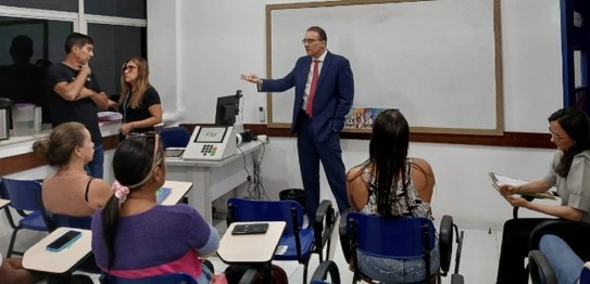 No centro da sala, o juiz veste um terno preto e conversa com os alunos, que estão sentados em c...