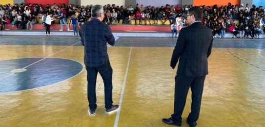 No centro da sala, o juiz veste um terno preto e conversa com os alunos, que estão sentados em c...