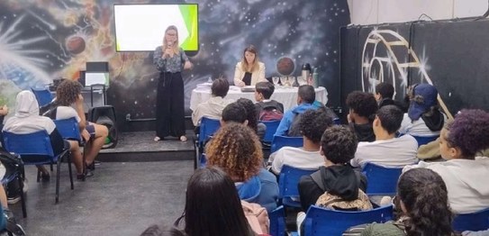 Descrição da imagem: O juiz veste um terno preto e segura um microfone em cima do palco. As(os) ...