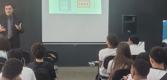 No centro da sala, o juiz veste um terno preto e conversa com os alunos, que estão sentados em c...