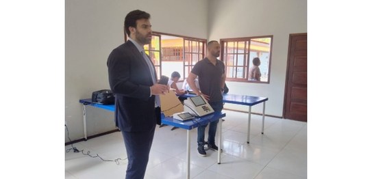 Alunos sentados assistem palestra feita por homem de terno, que aponta para duas urnas eletrônic...