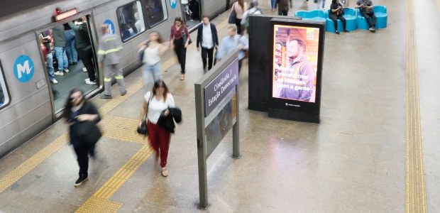 Vista do alto de uma estação de metrô em movimento. À esquerda, um trem passa em alta velocidade...