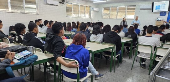 Descrição da imagem: O juiz veste um terno preto e segura um microfone em cima do palco. As(os) ...