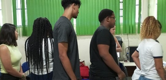 Descrição da imagem: Cinco alunos(as), três meninas e dois meninos, estão em pé e fazem fila par...