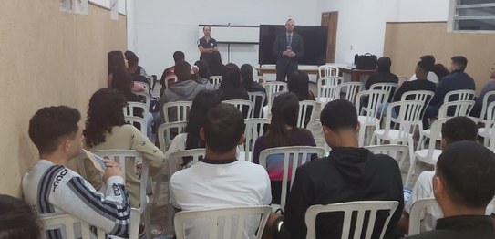 No centro da sala, o juiz veste um terno preto e conversa com os alunos, que estão sentados em c...