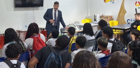 Descrição da imagem: Cinco alunos(as), três meninas e dois meninos, estão em pé e fazem fila par...